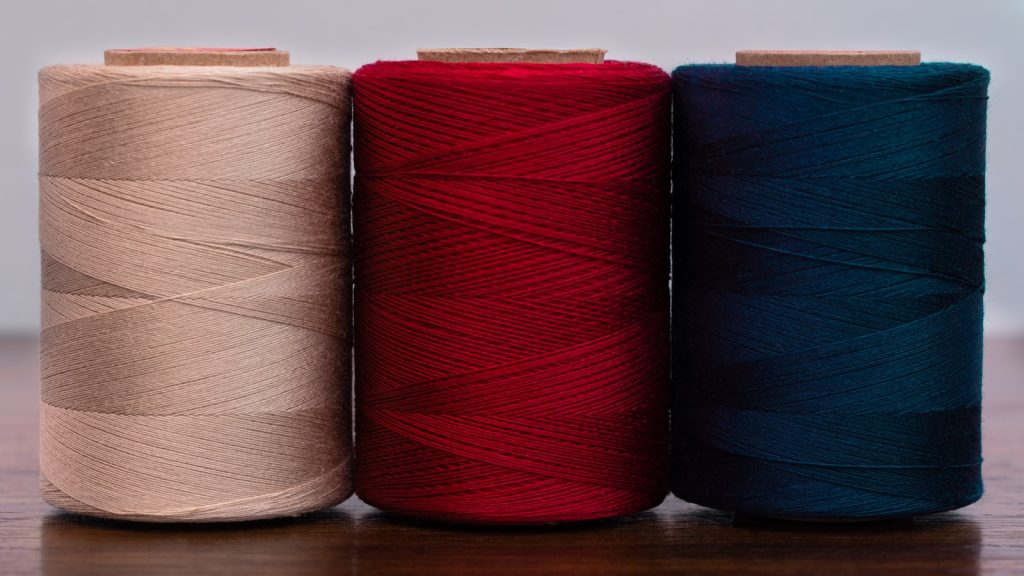 Trois bobines de fil beige, rouge et bleu alignées sur un plan de travail en bois