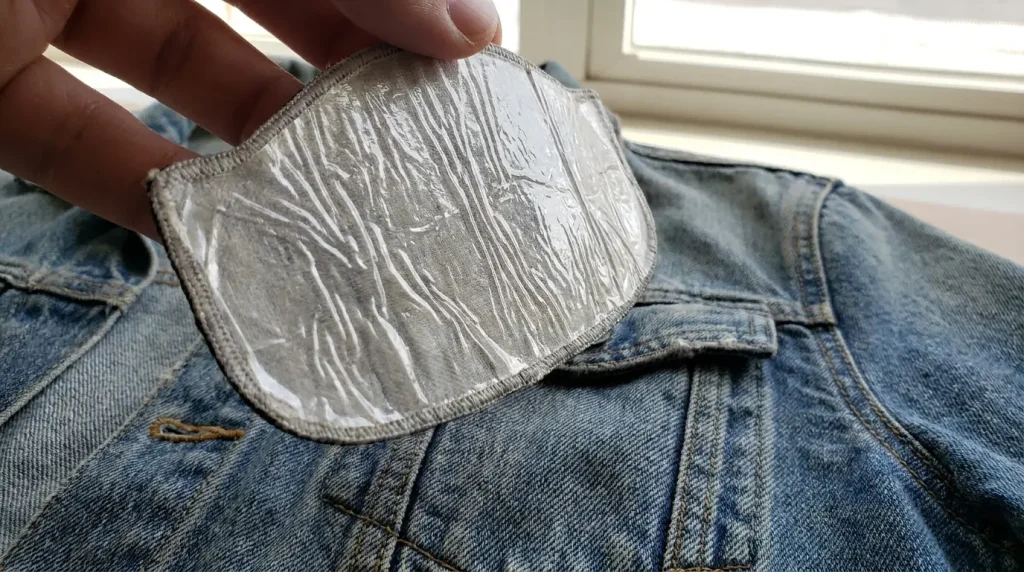 Envers d'un écusson brodé montrant sa fine pellicule de colle brillante, au-dessus d'un jean